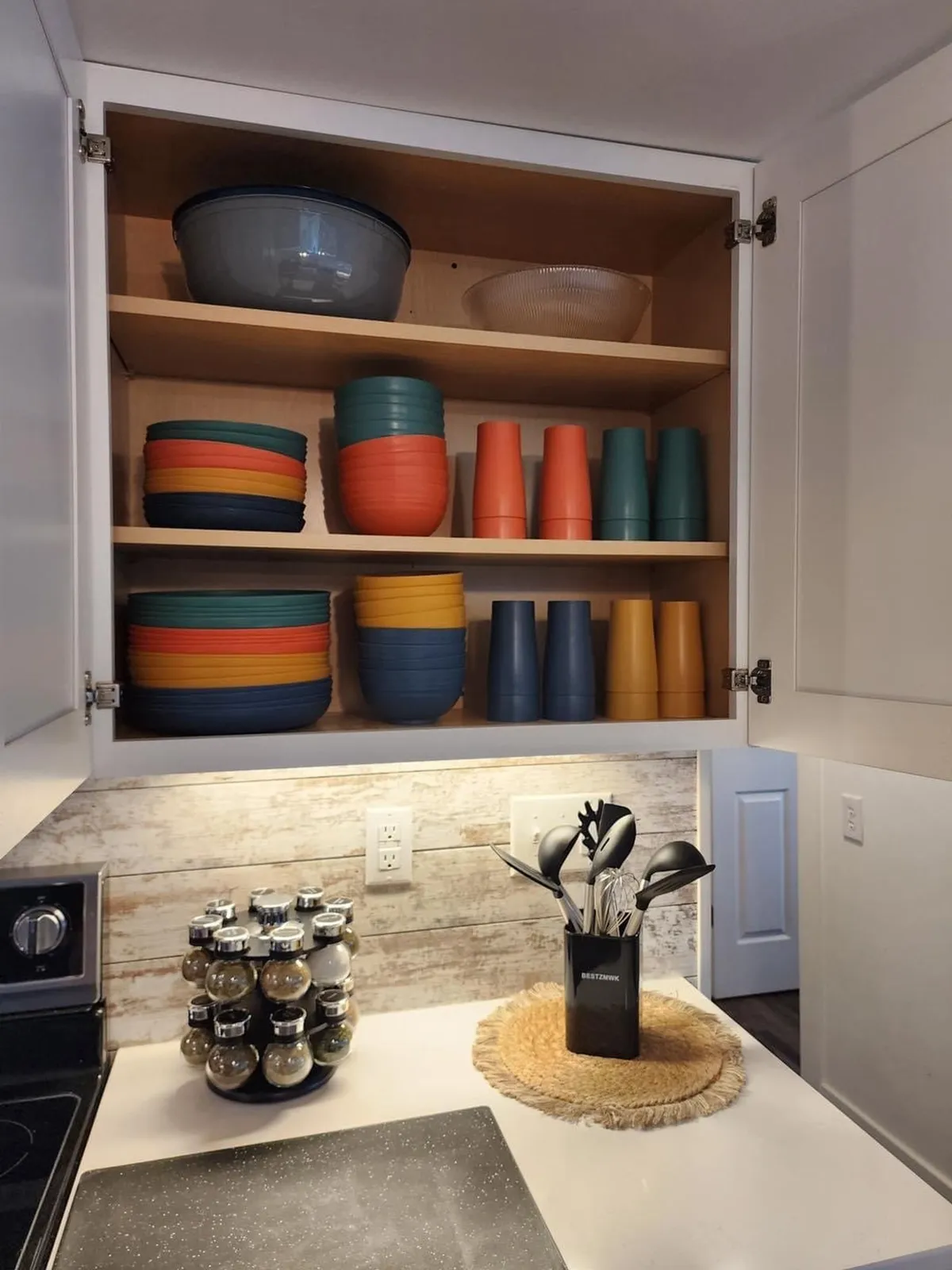 The image spotlights colorful dishware, a spice rack, and a utensil holder—key elements of a well-stocked kitchen. The cheerful arrangement of essentials visually supports the idea of the 'Pack Less Program' and the promise of gourmet amenities.