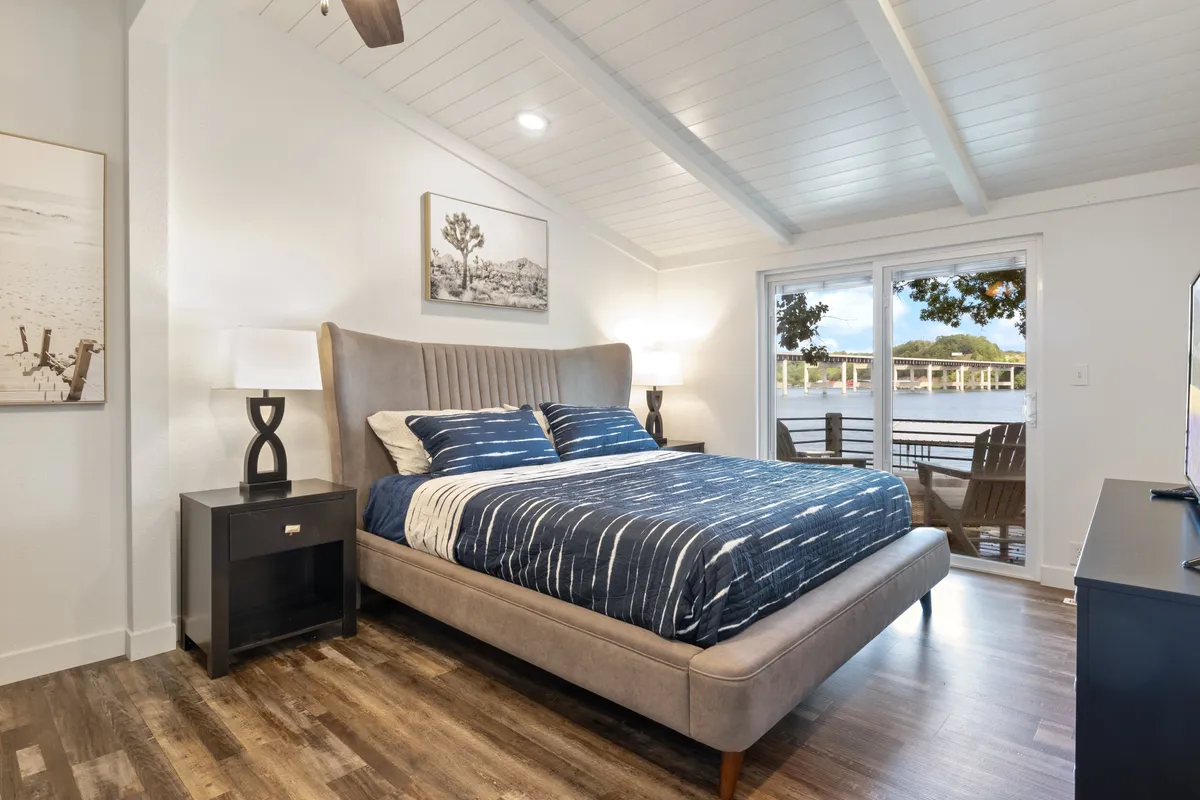 A tranquil bedroom with a large window offering glimpses of the water, a cozy bed made up with soft bedding, a stylish nightstand, and calming artwork above the bed. Natural light fills the room, highlighting the peaceful décor and inviting atmosphere.