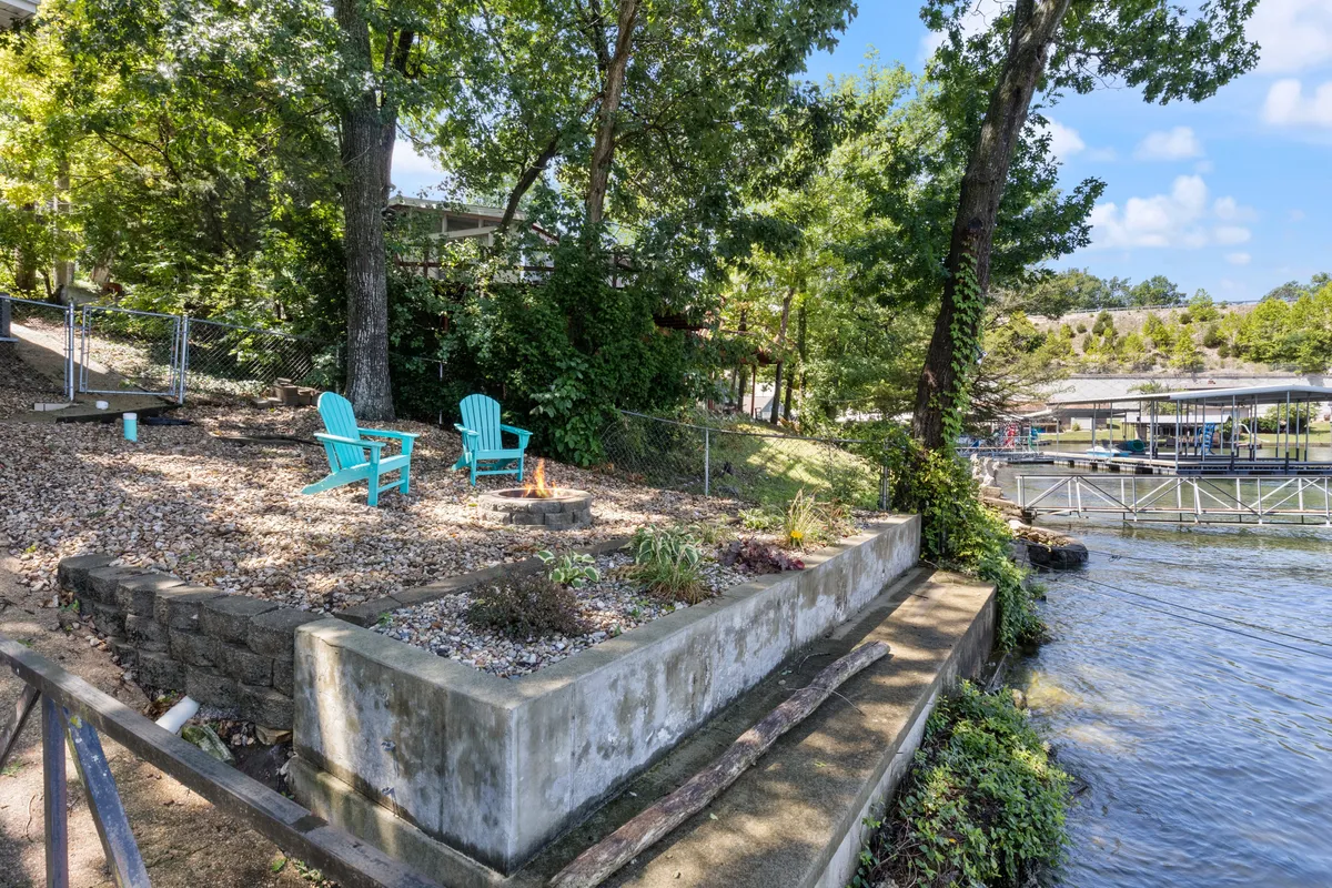 This photo shows two turquoise chairs arranged around a firepit area, set against a peaceful waterfront backdrop. The stone retaining wall and lush greenery frame the scene, making it ideal for evening gatherings, sharing stories, or enjoying s'mores as the sun sets over the water.