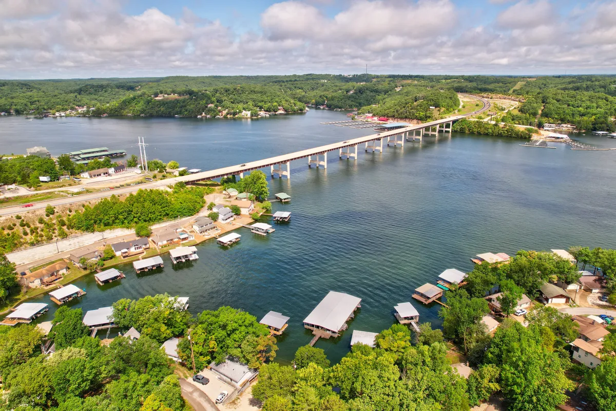 A sweeping view of the lake, bridge, waterfront homes, and abundant greenery, suggesting the broader context of local exploration. The vantage point includes water, trails, and neighboring attractions, evoking the spirit of discovery and the local area’s offerings.