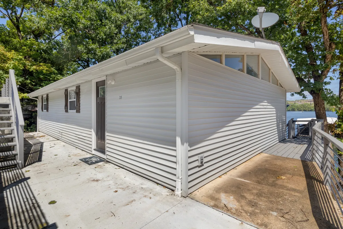 The image shows the exterior siding of the home, a spacious deck area, and surrounding trees. Steps lead up to the entrance, creating a welcoming first impression and a clear connection between indoor comfort and the scenic outdoors.