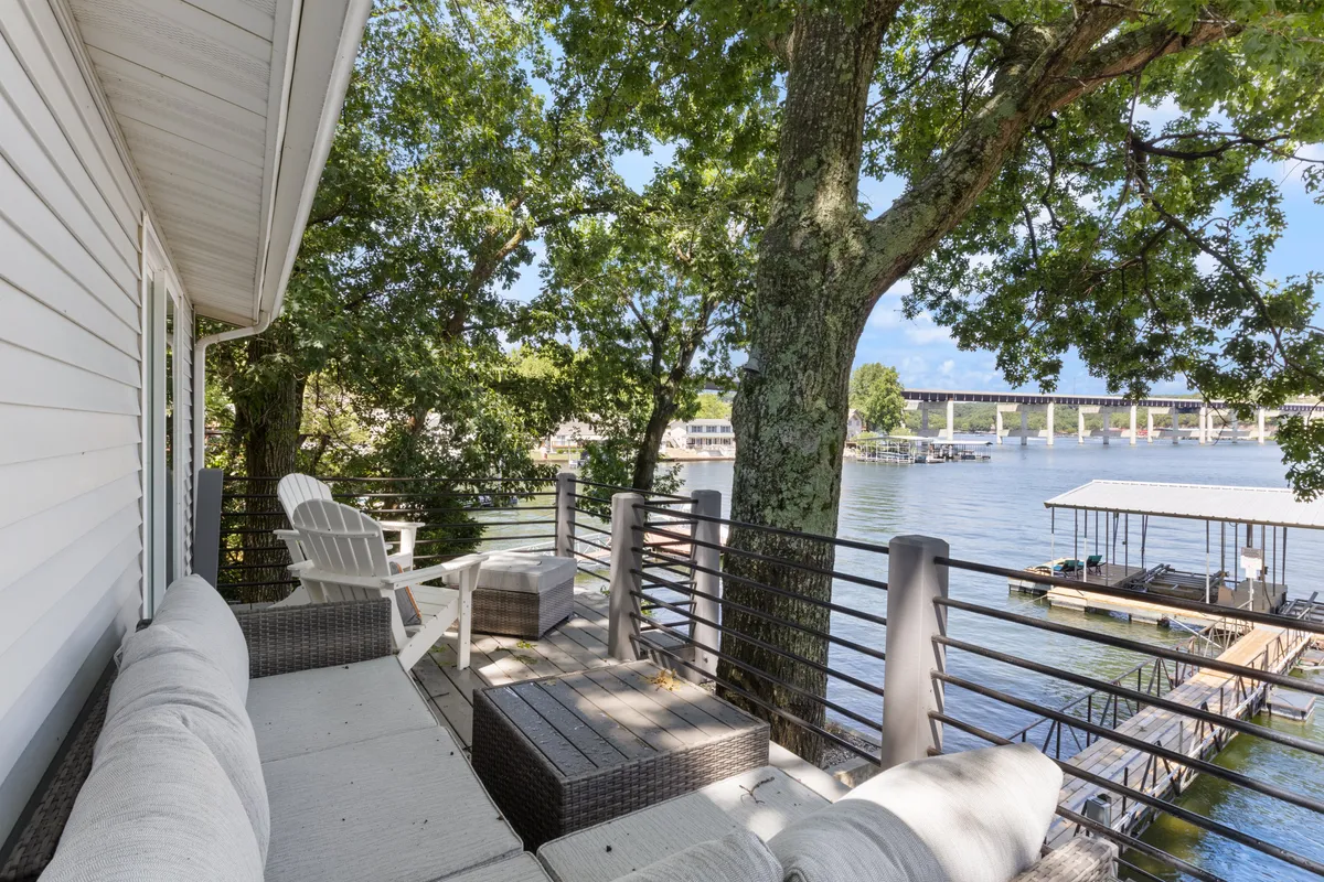 This image depicts a tranquil waterfront scene with outdoor seating arranged beneath a leafy tree canopy. The water is calm, and the setup invites guests to relax, socialize, or enjoy quiet moments while connecting with nature.