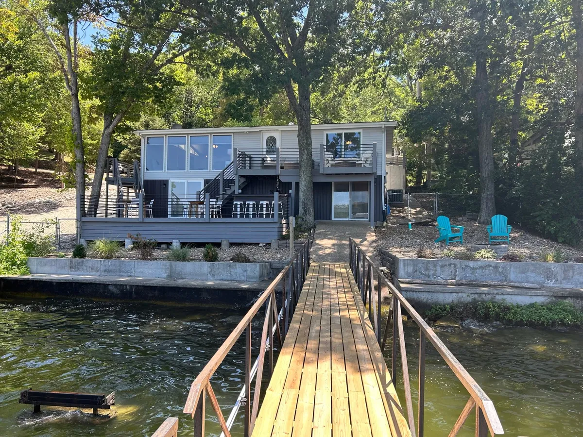 A striking shot showing the house with large lake-facing windows, a wooden dock leading up to the home, and vibrant blue Adirondack chairs set out for relaxing or entertaining. The entire scene is bathed in warm, golden-hour light.