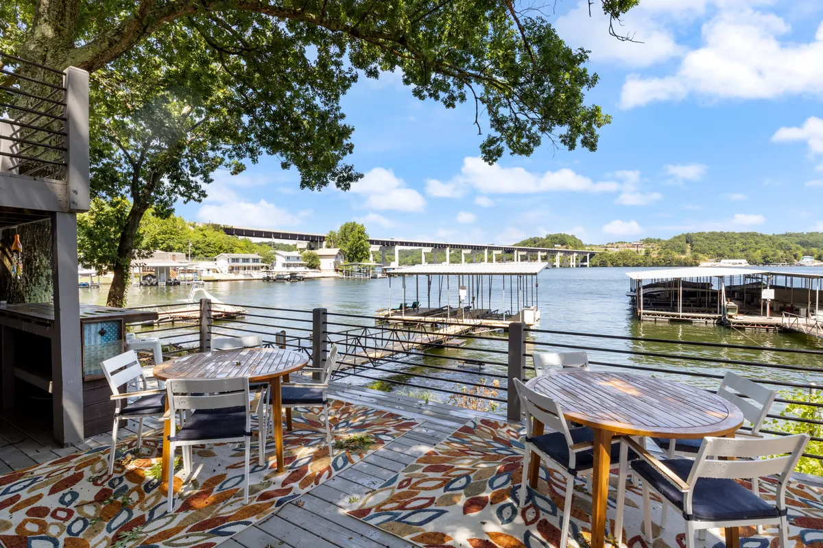 A spacious outdoor deck featuring elegant dining tables, a decorative rug, and a breathtaking view of the lake bordered by lush trees. The setup suggests a welcoming environment for alfresco meals and relaxed conversation.