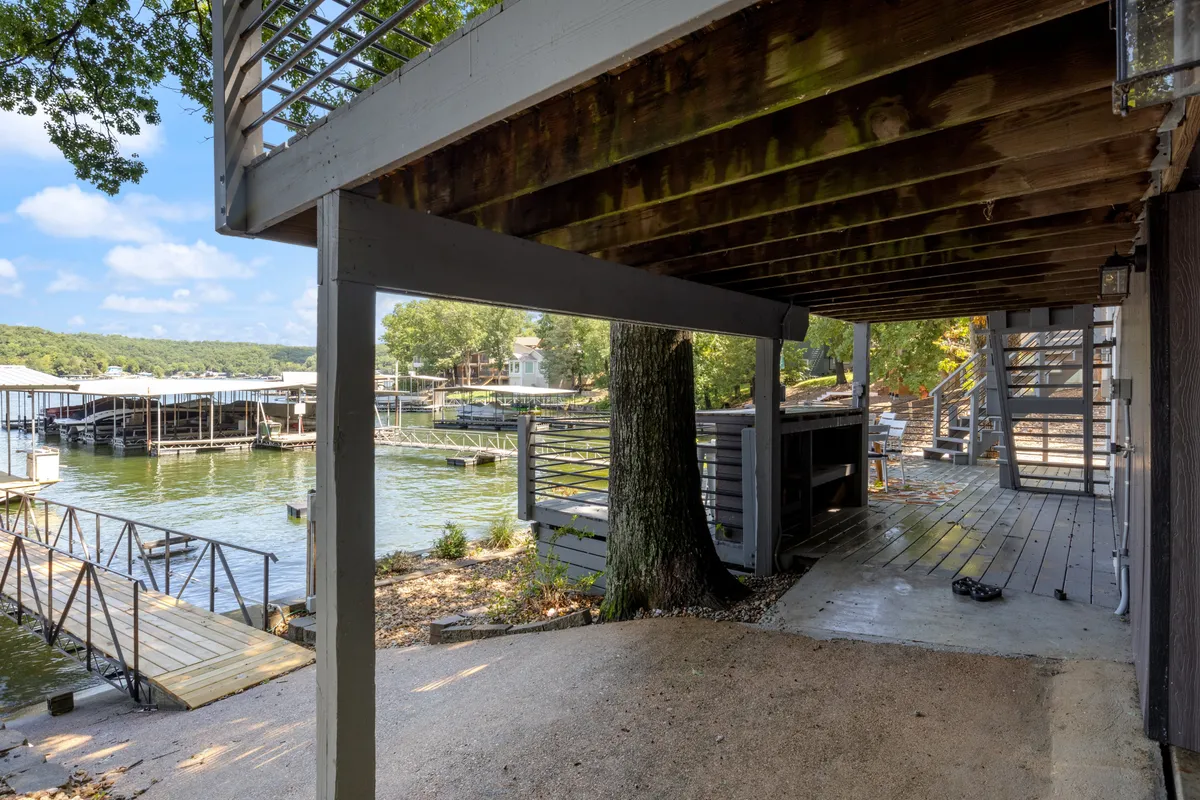 A wide view of a private dock extending over calm lake waters, with a tree providing gentle shade and the deck area visible above. The scene hints at activities like boating, fishing, and sunset relaxation, while the tranquility of the setting suggests privacy and exclusivity.
