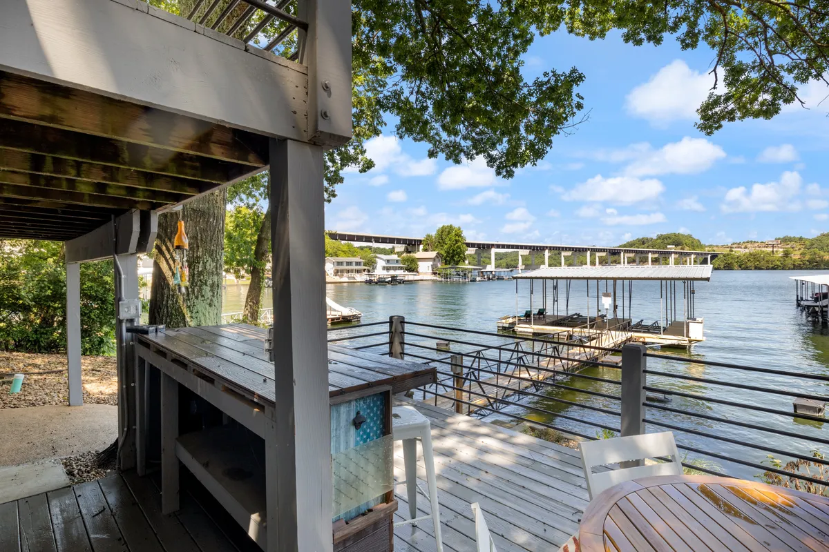 A serene lakeside setting with outdoor seating on a dock, overlooking still water and a canopy of trees. The space invites relaxation, conversation, or enjoying a morning coffee with tranquil water views. The arrangement suggests access to boating, fishing, or evening gatherings by the lake.
