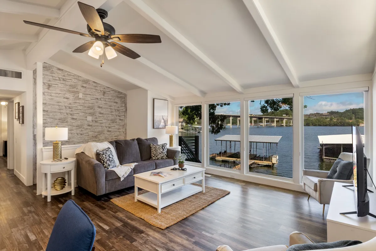 A contemporary living room with a comfortable gray sofa, decorative throw pillows, a wooden coffee table, and expansive windows offering a direct water view. Sunlight streams in, illuminating the tranquil space designed for both relaxation and entertaining guests.