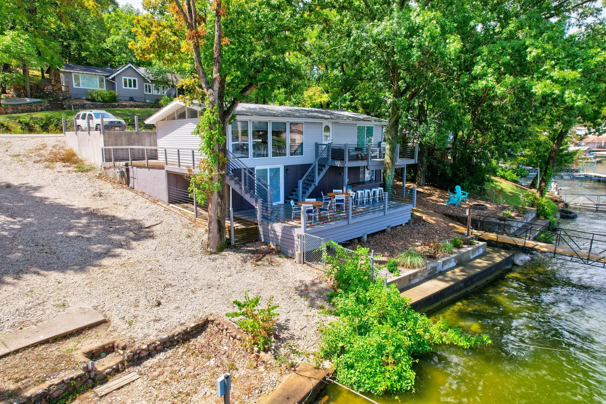 An inviting deck with comfortable seating overlooks the lake, surrounded by greenery. Stairs lead down to the water, showing easy access for boating, fishing, or swimming. The setting is ideal for morning coffee or evening barbecues while enjoying views from both upper and lower decks.