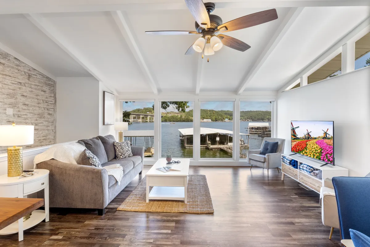 The image showcases a spacious, inviting living area with a plush, comfortable sofa facing large windows that frame stunning lake views. Natural light pours in, creating a serene ambiance perfect for relaxing or gathering with loved ones. A TV mounted on the wall hints at cozy movie nights, while the open layout suggests space for group socializing.